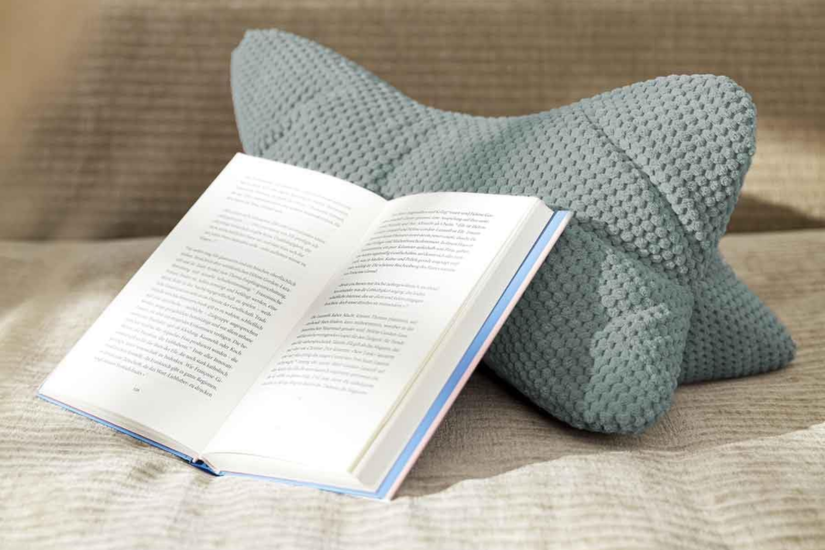 Gemütliche Szene mit einem offenen Buch, das an ein strukturiertes, graublaues Nackenkissen gelehnt ist – platziert auf einem beigefarbenen Sofa oder Bett.